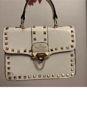 Vit Valentino Garavani handväska med nitar - Snygg vit handväska från Valentino Garavani i läder med guldiga nitar runt kanterna och på handtaget. Väskan har ett elegant lås framtill med kedjedetalj och ett stadigt handtag i svart och vitt. Perfekt för dig som gillar lyxiga accessoarer med attityd.
