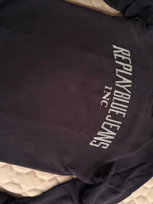 Svart sweatshirt från Replay - Cool svart sweatshirt från Replay med trycket 'REPLAY BLUE JEANS INC.' i vitt framtill. Tröjan har rund halsringning och långa ärmar. Perfekt för en avslappnad och trendig look.