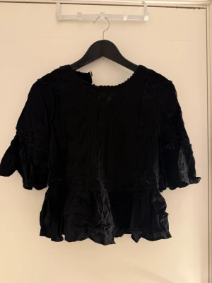 Svart volangblus med spetsdetaljer - Trendig svart blus med volangkant och fina spetsdetaljer. Blusen har rund halsringning och korta puffärmar som ger en romantisk vibe. Perfekt att styla med både jeans och kjol för en cool look.