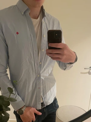 Ljusblå skjorta från Morris - Säljer en ljusblå skjorta från Morris med klassisk button-down krage och liten röd broderad logga på bröstet. Skjortan är tillverkad i bomull och har vita knappar framtill. Perfekt för en clean och stilren look.