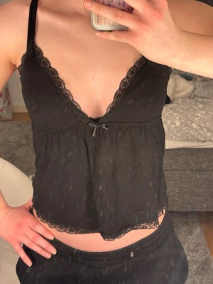 Svart linne från Nelly - Svart linne med tunna axelband och v-ringning. Linnet har spetsdetaljer längs urringningen och nederkanten samt en liten rosett framtill. 