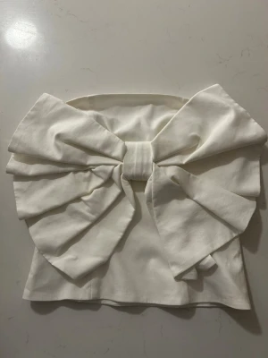 Vit axelbandslös topp med stor rosett - Supersnygg axelbandslös topp i vitt med en stor rosett framtill. Toppen har bandeau-modell och dragkedja i sidan för enkel på- och avtagning. Perfekt för dig som vill sticka ut och addera något extra till din outfit.