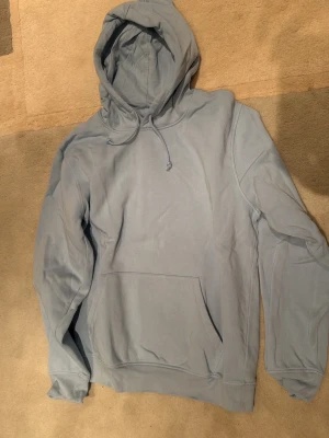 Ljusblå hoodie med ficka - En stilren ljusblå hoodie med huva och dragsko. Hoodien har en stor magficka framtill och ribbade muddar vid ärmslut och nederkant. Tillverkad i mjukt bomullsliknande material som känns skönt mot huden. Perfekt för en avslappnad och trendig look.