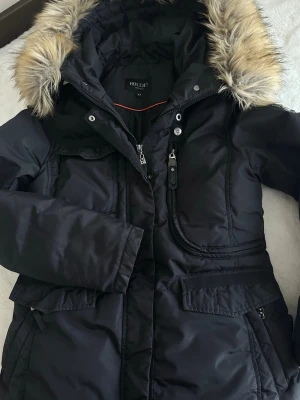 Svart Hollie’s jacka  - Snygg svart parkasjacka från Hollies med avtagbar päls. Jackan har flera fickor med dragkedja och knappar, justerbar midja och varm vaddering. Perfekt för kalla dagar och riktigt skön att bära. Nypris 4000kr! Inga hål inga fläckar💕