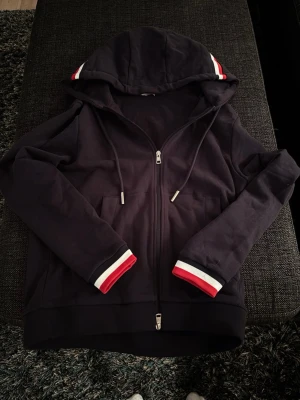 Svart hoodie från Moncler med dragkedja - Snygg svart hoodie från Moncler med dragkedja framtill och huva med dragsko. Ärmsluten och huvan har stilrena ränder i vitt och rött. Tillverkad i mjukt bomullsmaterial som känns skönt mot huden. Perfekt för en avslappnad och trendig look.