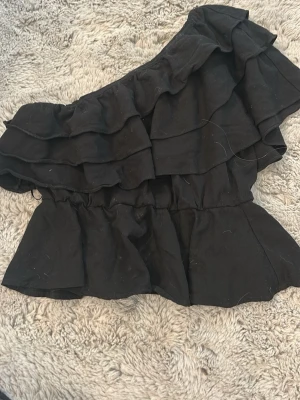 Svart oneshoulder volangblus Vero Moda - Snygg svart oneshoulder blus från Vero Moda med flera lager volanger. Tillverkad i mjuk bomull och har en croppad passform som ger en trendig look. Perfekt för dig som vill sticka ut med en cool och unik topp.