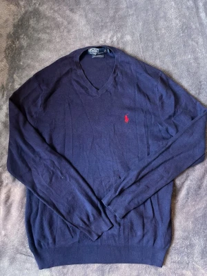  tröja från  Polo Ralph Lauren - Mörkblå v-ringad tröja från Polo Ralph Lauren med den klassiska röda logotypen broderad på bröstet. Tröjan har långa ärmar och är tillverkad i ett tunt, mjukt material som känns skönt mot huden. Perfekt för en stilren och avslappnad look.