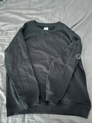 Svart sweatshirt från C.P. Company - Snygg svart sweatshirt från C.P. Company med rund halsringning och ribbade muddar. Tröjan har en ikonisk linsdetalj på ena ärmen och är tillverkad i mjukt bomullsmaterial. Perfekt för en clean och stilren look.