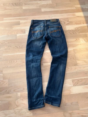 Mörkblå jeans från Nudie Jeans - Säljer ett par mörkblå jeans från Nudie Jeans med klassisk femficksmodell och orangea sömmar. Jeansen har raka ben och snygg slitning framtill och baktill. Tillverkade i denim med en autentisk tvättad look. Perfekta för en avslappnad och trendig stil.