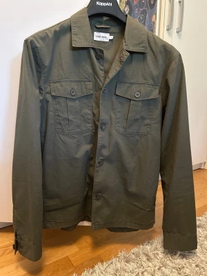 Olivgrön overshirt från East West - Stilren olivgrön overshirt från East West med klassisk krage och två bröstfickor med lock och knapp. Långärmad modell med knäppning framtill och vid ärmslut. Perfekt för lager-på-lager och enkel att matcha med olika outfits.