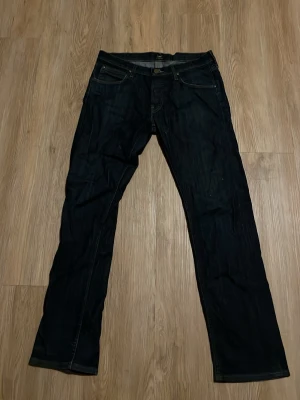 Mörkblå raka jeans från Lee - Säljer ett par mörkblå jeans från Lee med klassisk rak passform och fem fickor. Jeansen har kontrastsömmar och Lee-logga på bakfickan samt läderpatch i midjan. Tillverkade i slitstark denim som ger en tidlös look.