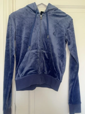 Blå velour hoodie från Juicy Couture - Mjuk och lyxig blå velour hoodie från Juicy Couture med dragkedja framtill, fickor och justerbar huva med snören. Diskret broderad logga på bröstet och breda muddar vid ärmar och midja. Perfekt för en chill och trendig look.