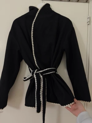 Svart kappa med vit kant och knytskärp - Stilren svart kappa med vit dekorativ kant längs krage, öppning och nederkant. Kappan har ett matchande knytskärp i midjan och raka ärmar. Perfekt för dig som gillar minimalistisk men ändå unik design. Materialet ser ut att vara mjukt tyg, troligen bomull eller polyester.