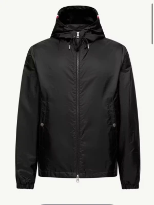 Svart vindjacka från Moncler - Stilren svart vindjacka från Moncler med huva och dragkedja framtill. Jackan har två sidofickor med knappstängning och diskret Moncler-logga på ärmen. Materialet är lätt och vattenavvisande, perfekt för blåsiga dagar. Huva med snörning och elastiska muddar. Är ute efter byte mot en större storlek M eller L  går att köpa också. Jackan är knappt använd
