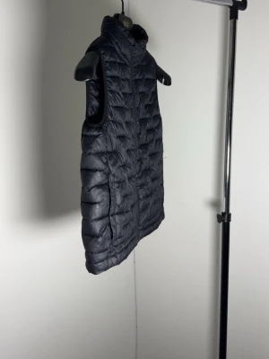 Svart quiltad dunväst med dragkedja - Stilren svart dunväst med quiltat mönster och hög krage. Västen har hel dragkedja framtill och en klassisk, rak passform. Perfekt att slänga över en hoodie eller tröja för extra värme. Enkel och tidlös design utan synligt märke.