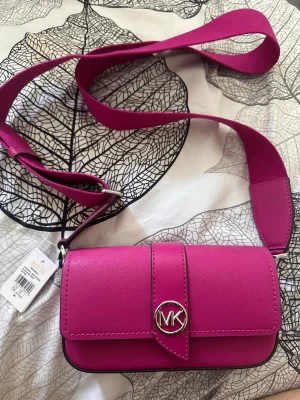 Rosa axelväska från Michael Kors - Snygg axelväska i stark rosa färg från Michael Kors med bred axelrem och silverfärgat MK-spänne framtill. 