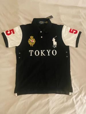 Svart pikétröja Tokyo Polo Ralph Lauren - Svart pikétröja från Polo Ralph Lauren med vita ärmar och röda siffror på axlarna. Stort Tokyo-tryck framtill, broderad logga och emblem i gult och vitt. Klassisk krage och knappar vid halsen. Perfekt för en sportig och stilren look.