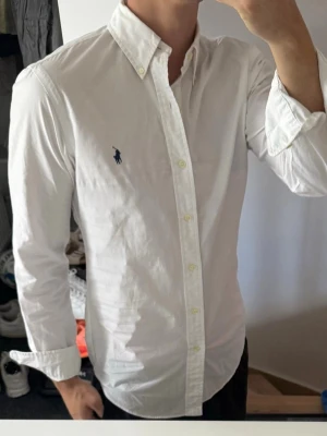 Vit skjorta från Ralph Lauren - Klassisk vit skjorta från Ralph Lauren med button-down krage och broderad marinblå logga på bröstet. Skjortan har långa ärmar, knappar framtill och är tillverkad i bomull för en bekväm och stilren look.