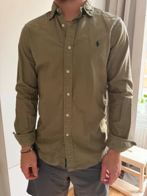 Olivgrön skjorta från Ralph Lauren - Klassisk olivgrön skjorta från Ralph Lauren med lång ärm och knappar framtill. Skjortan har en liten mörkblå logga broderad på bröstet och är tillverkad i mjuk bomull. Perfekt till jeans eller chinos för en avslappnad men snygg look.