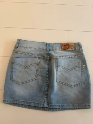 Ljusblå jeanskjol från Dear Marks - Säljer en ljusblå jeanskjol från Dear Marks med klassisk femficksdesign och bälteshällor. Kjolen har en kort passform och är tillverkad i mjukt denimtyg. Perfekt för dig som gillar en enkel och tidlös stil.