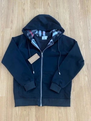 Burberry Hoodie - Snygg svart hoodie från Burberry med dragkedja och klassiskt rutigt foder i huvan. Hoodien har dragsko i huvan, två fickor fram och diskret Burberry-logga broderad på bröstet. Perfekt för dig som gillar stilrena och exklusiva plagg.