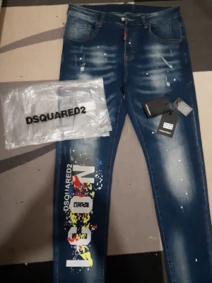 Blå Dsquared2 jeans med tryck - Säljer ett par blå Dsquared2 jeans med coolt färgstarkt tryck och texten ICON på vänster ben. Jeansen har slitningar, vita färgstänk och klassisk femficksmodell. Materialet är denim och passformen är smal. Perfekta för dig som vill sticka ut med din stil.