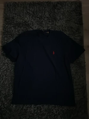 Mörkblå t-shirt från Ralph Lauren - Snygg mörkblå t-shirt från Ralph Lauren med klassisk röd logga broderad på bröstet. T-shirten har korta ärmar och en enkel, stilren design som passar till det mesta. Perfekt basplagg för garderoben.