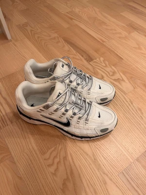 Nike P6000 - Snygga vita Nike sneakers med silverfärgade detaljer och svarta loggor. Skorna har en sportig design med mesh och syntetmaterial, samt en låg siluett och rejäla snören. Perfekta för dig som gillar streetwear och vill ha en clean look.