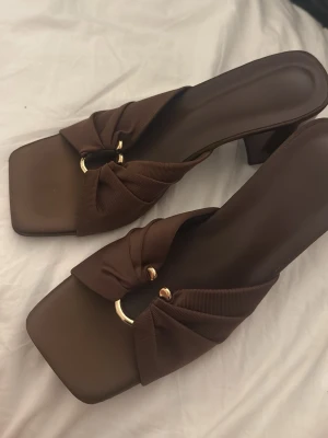 Bruna slip-in sandaler med klack - Stilrena bruna sandaler med fyrkantig tå och bred klack. Ovandelen har en twistad design i tyg med guldfärgade ringdetaljer. Perfekta för dig som gillar trendiga och bekväma skor med en modern touch.