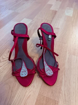 Röda strassprydda pumps med remmar - Snygga röda pumps med smala remmar och öppen tå. Skorna har en elegant klack och är dekorerade med glittrande strass framtill. Perfekta för att sticka ut och ge outfiten extra bling. Materialet ser ut att vara satinliknande tyg. Jag har aldrig använt de därför säljer jag. Köpt på vinted
