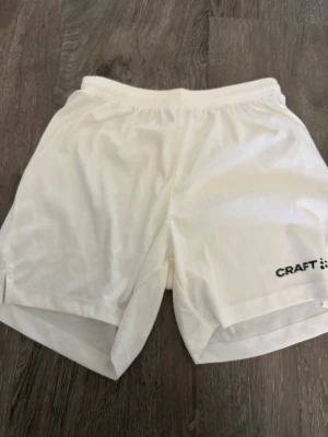 Vita träningsshorts från Craft - Säljer ett par vita träningsshorts från Craft med elastisk midja och diskret logga på vänster ben. Shortsen har en klassisk passform och är tillverkade i ett lätt och funktionellt syntetmaterial som passar perfekt för sport och aktivitet.