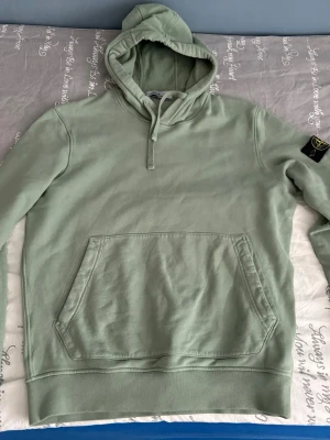 Grön hoodie från Stone Island - Snygg ljusgrön hoodie från Stone Island med klassisk patch på vänster ärm. Hoodien har huva med dragsko och stor magficka framtill. Tillverkad i mjuk bomull för skön känsla och avslappnad stil.
