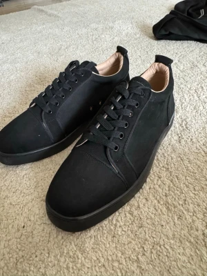 Christian Louboutin skor - Stilrena svarta sneakers i mocka från Christian Louboutin med klassisk röd sula och svarta skosnören. Låg modell med diskreta detaljer och beige innerfoder. Perfekta för dig som vill ha en clean och exklusiv look.