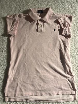 Ljusrosa pikétröja från Polo Ralph Lauren - Säljer en ljusrosa pikétröja från Polo Ralph Lauren med klassisk krage och två knappar framtill. Tröjan har korta ärmar och den ikoniska broderade loggan på bröstet. Perfekt för en clean och stilren look.