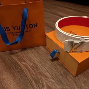 Vitt L bälte - Snyggt vitt skärp  med klassiskt L*-spänne i silver och präglat monogrammönster. Insidan är röd för en extra touch. Perfekt accessoar för att lyfta din outfit.