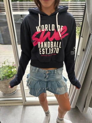 Svart hoodie Partille Cup - Snygg svart cropped hoodie från Partille Cup med vit och rosa text framtill. Hoodien har huva med vita snören och tryck på ena ärmen. Perfekt för dig som gillar sportig stil och vill sticka ut med coolt tryck.