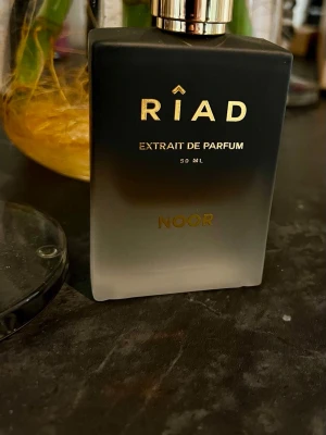 Riad Noor Extrait de Parfum 50 ml - Riad Noor Extrait de Parfum är en lyxig parfym i en elegant svart och frostad glasflaska med gulddetaljer. Flaskan rymmer 50 ml och kommer i en snygg röd kartong med guldfärgad text. Perfekt för dig som gillar exklusiva dofter.