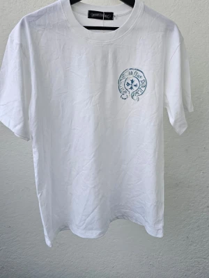 Vit t-shirt från Chrome Hearts - Vit t-shirt från Chrome Hearts med blått tryck på bröstet och stort grafiskt tryck på ryggen. Klassisk rund hals och korta ärmar. Tillverkad i mjuk bomull som känns skön mot huden. Perfekt för dig som gillar streetwear och snygga detaljer.