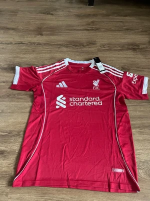 Liverpool matchtröja röd Adidas - Säljer en röd Liverpool matchtröja från Adidas med vita detaljer och klubbmärke på bröstet. Tröjan har korta ärmar, rund hals och tryckta loggor från Standard Chartered och Expedia. Perfekt för fotbollsfans som vill visa sitt stöd.