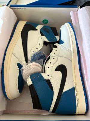 Nike Air Jordan 1 High OG blå/vit - Snygga Nike Air Jordan 1 High OG sneakers i blått, vitt och svart. Skorna har klassisk hög siluett, svart swoosh och blå detaljer på tå och häl. Ovandelen är i läder med perforerad tåbox och extra skosnören medföljer. Perfekta för dig som gillar streetwear och ikoniska sneakers.