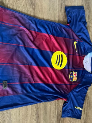Barcelona matchtröja Nike blå/röd - Säljer en Barcelona matchtröja från Nike i klassiska blå och röda vertikala ränder. Tröjan har gula logotyper, klubbmärke och sponsortryck på bröstet samt UNHCR-tryck på ryggen. Tillverkad i lätt och ventilerande funktionsmaterial, perfekt för fotbollsträning.