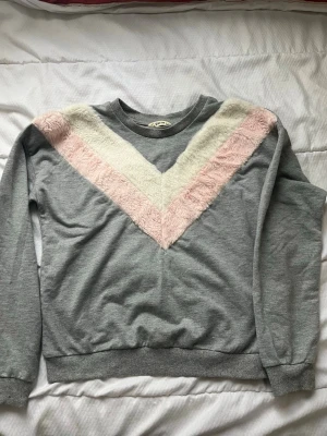 Grått loungewear-set med fluffdetaljer - Mysigt loungewear-set i mjuk grå bomullsblandning. Tröjan har lång ärm och fluffiga detaljer i vitt och ljusrosa i ett V-mönster över bröstet. Byxorna har resårmidja med dragsko och raka ben. Perfekt för chill dagar hemma. Den är mycket använd med defekter som mycket fläckar.