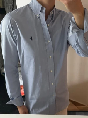 Ljusblå skjorta från Polo Ralph Lauren - Klassisk ljusblå skjorta från Polo Ralph Lauren med button-down krage och broderad logga på bröstet. Skjortan har långa ärmar, vita knappar och är tillverkad i bomull. Perfekt för en clean och stilren look. storlek 14 1/2 vilket motsvarar m ungefär 