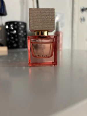 Rituals parfym rosa - Säljer en snygg parfym från Rituals i en fyrkantig rosa glasflaska med en unik träliknande kork och gulddetaljer. Flaskan har en modern och stilren design som känns lyxig och trendig. Perfekt för dig som gillar exklusiva dofter och vill ha en fin flaska på hyllan. Den är i fint skick men använd!💕