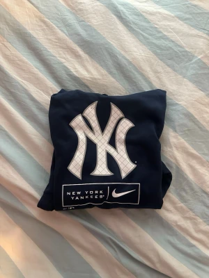 NY Yankees Nike Hoodie - Snygg mörkblå hoodie med stor New York Yankees-logga på bröstet och Nike-logga under. Klassisk känguruficka framtill och justerbar huva med dragsko. Perfekt för dig som gillar sportig streetwear och vill sticka ut med en ikonisk look.