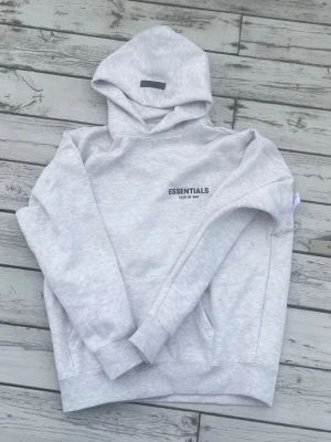 Grå Essentials Fear of God hoodie - Snygg ljusgrå hoodie från Essentials Fear of God med svart logga tryckt på bröstet och ryggen. Klassisk passform med huva och ribbade muddar vid ärmar och midja. Perfekt för en chill och stilren look. Tillverkad i mjukt bomullsmaterial.