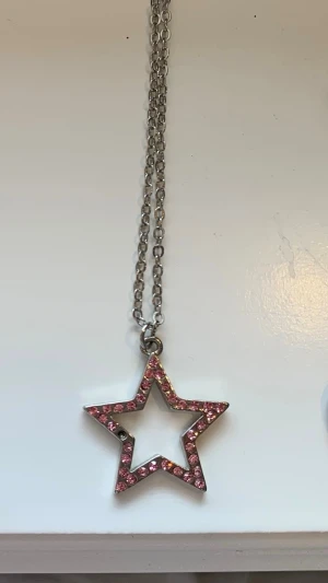 Halsband med stjärnhänge och rosa stenar - Snyggt halsband med en silverfärgad kedja och ett stjärnformat hänge dekorerat med glittrande rosa stenar. Perfekt för dig som gillar statement-smycken och vill addera lite färg till din outfit. Kedjan är av metall och hänget har en öppen design.