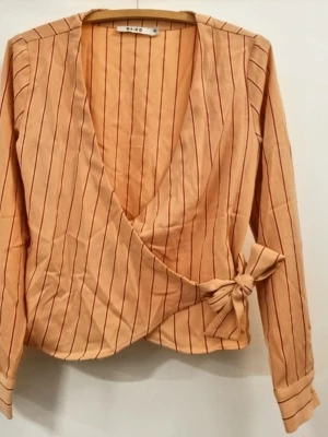 Beige omlottblus med ränder från NA-KD - Trendig beige omlottblus från NA-KD med tunna röda vertikala ränder och knytband i sidan. Blusen har djup V-ringning och långa ärmar med knappdetalj vid ärmslut. Perfekt för en stilren och modern look.