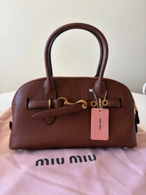 Brun handväska från Miu Miu i läder - Snygg brun handväska från Miu Miu i äkta läder med guldfärgade metalldetaljer och dubbla handtag. Väskan har en stilren, rundad form och kommer med original dustbag och äkthetskort. Perfekt accessoar för att lyfta din outfit.