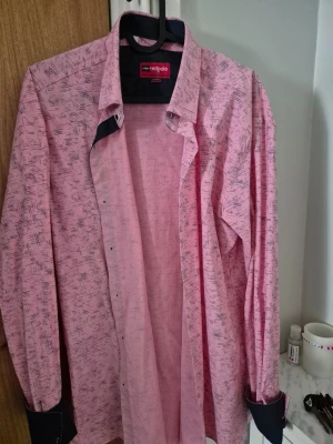 Rosa skjorta från Redpolo slim fit - Snygg rosa skjorta från Redpolo med diskret abstrakt mönster i svart. Slim fit-modell med långa ärmar och mörkblå detaljer på insidan av kragen och manschetterna. Perfekt för dig som gillar en modern och stilren look.